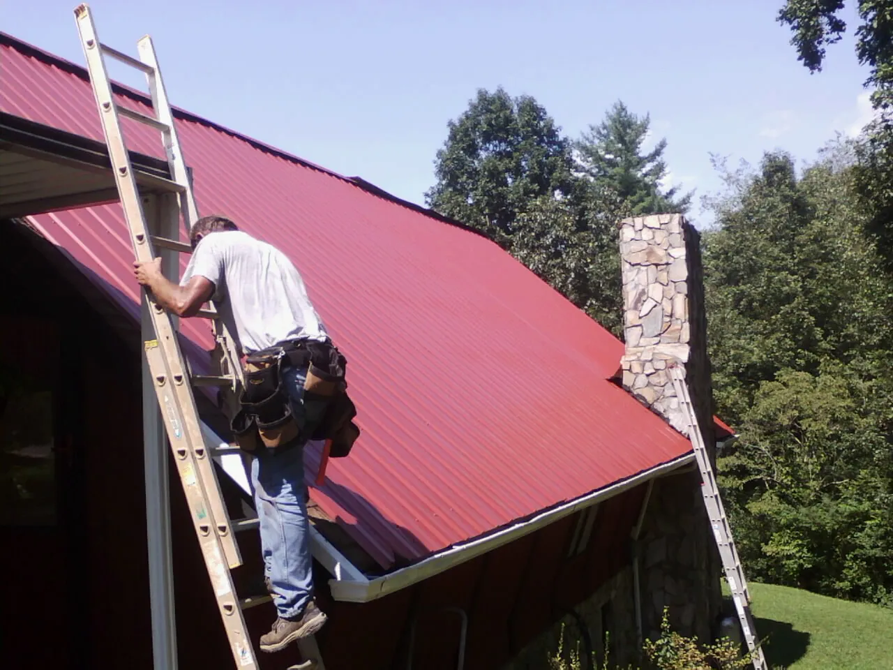 Roofer on a ladder beside a red metal roof and stone chimney — Soffit & Fascia Repair by Roofing Brothers in Juniata